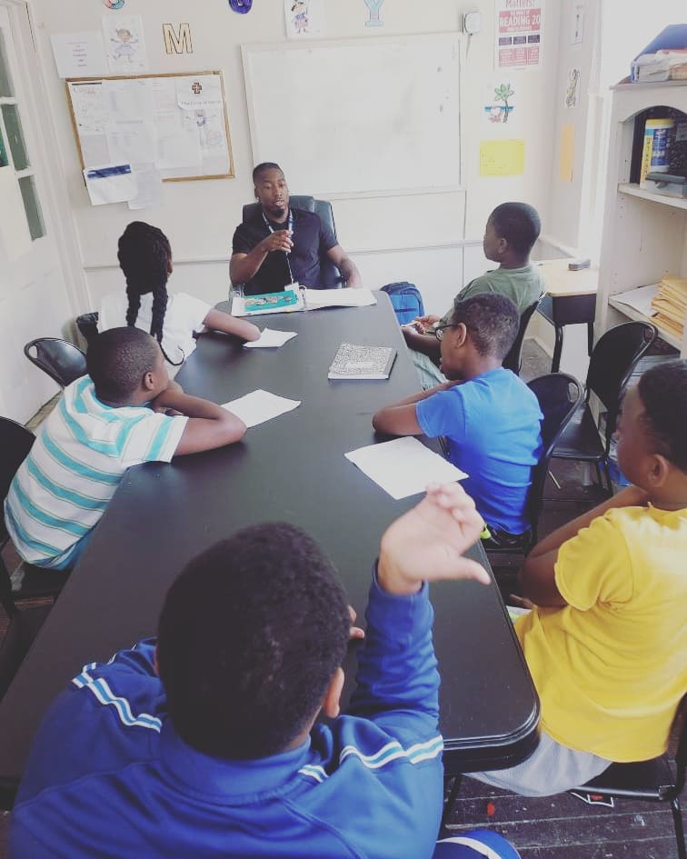 Six African American children sit around a black table in a classroom. An adult African American male is sitting at the head of the table.