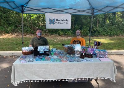 A man and a woman selling masks under a canopy. A sign hangs in the back with a blue butterfly and the text "Blue Butterfly by Carrie Marie."