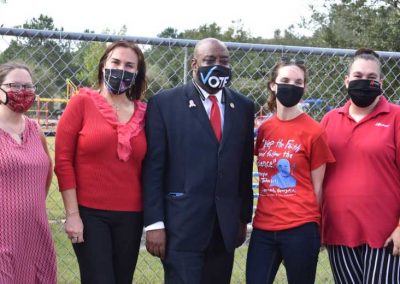 Five adults standing in front of a chain-link fence. The four women are wearing red shirts and dresses and the one man is wearing a dark suit