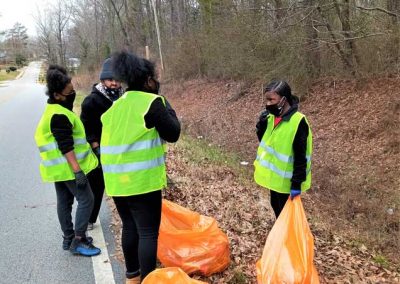 Four people, two of whom are wearing yellow vests, put trash from alongside a road into orange garbage bags.